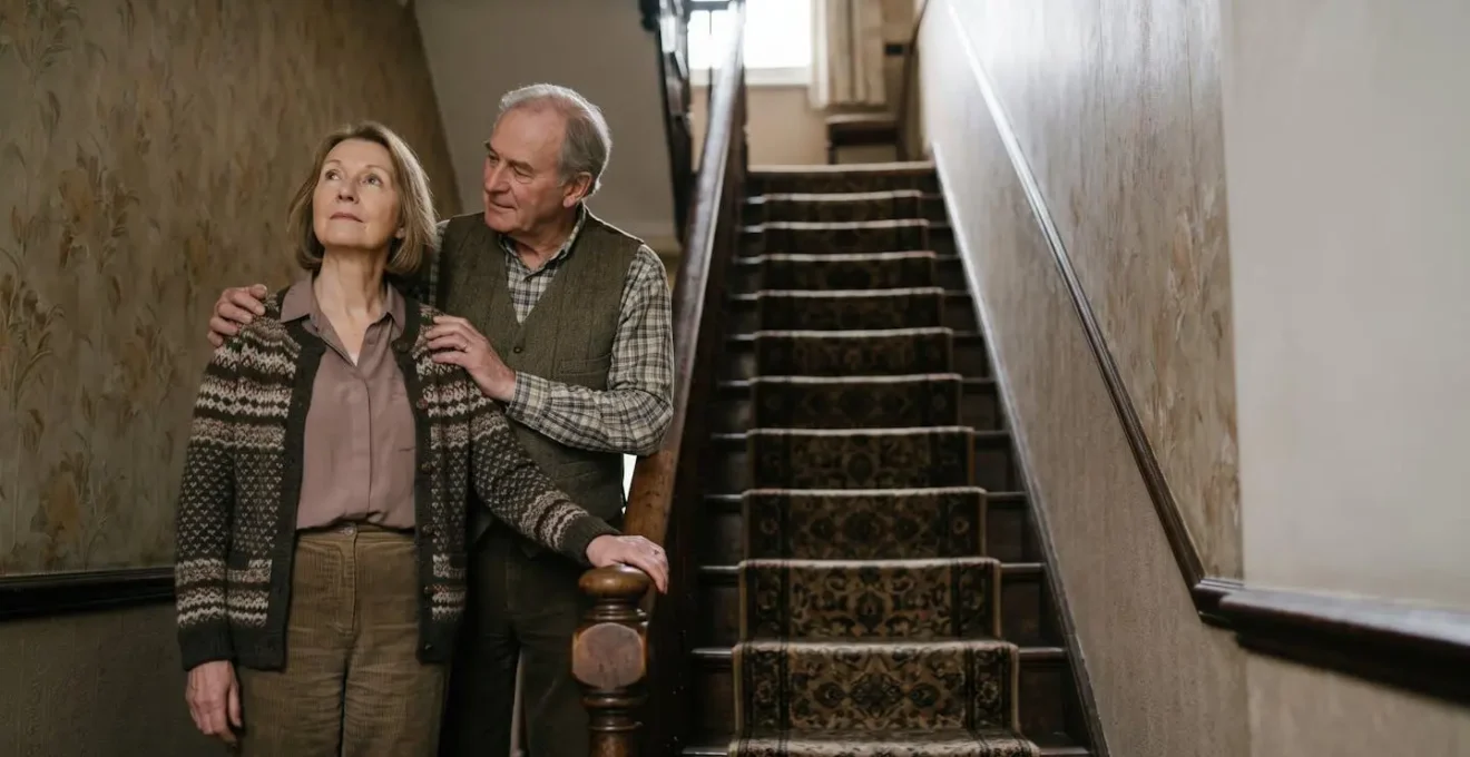 Elderly couple thoughtfully standing at the bottom of a Victorian terraced house staircase, contemplating mobility solutions