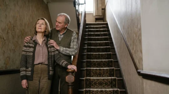 Elderly couple thoughtfully standing at the bottom of a Victorian terraced house staircase, contemplating mobility solutions